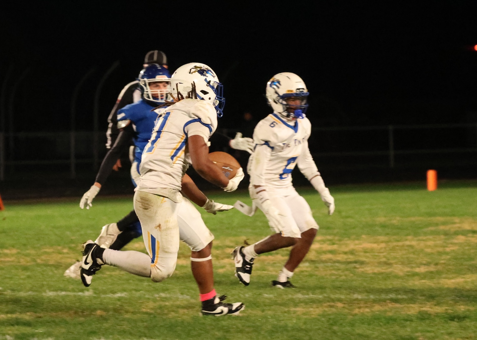 A football player in a white uniform carrying the ball runs on a night game, with teammates and a pursuing blue-clad defender nearby on a grass field.