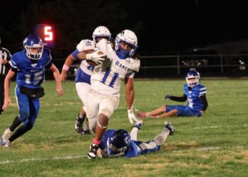 PY8A2292 1 | Press Pass Sports White-number-11 football player runs with the ball, evading blue defenders on a nighttime field as a tackler lies on the grass nearby.