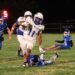 White-number-11 football player runs with the ball, evading blue defenders on a nighttime field as a tackler lies on the grass nearby.