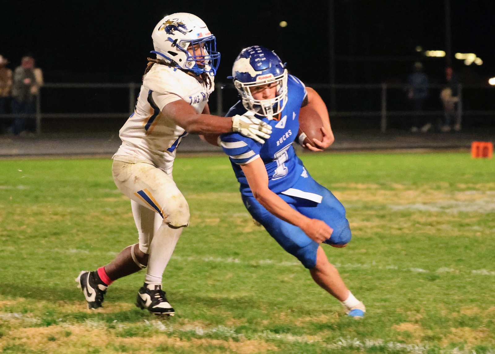 Football players chase the ball on a night game; blue jersey runner keeps possession while tackled by a white-jersey defender.