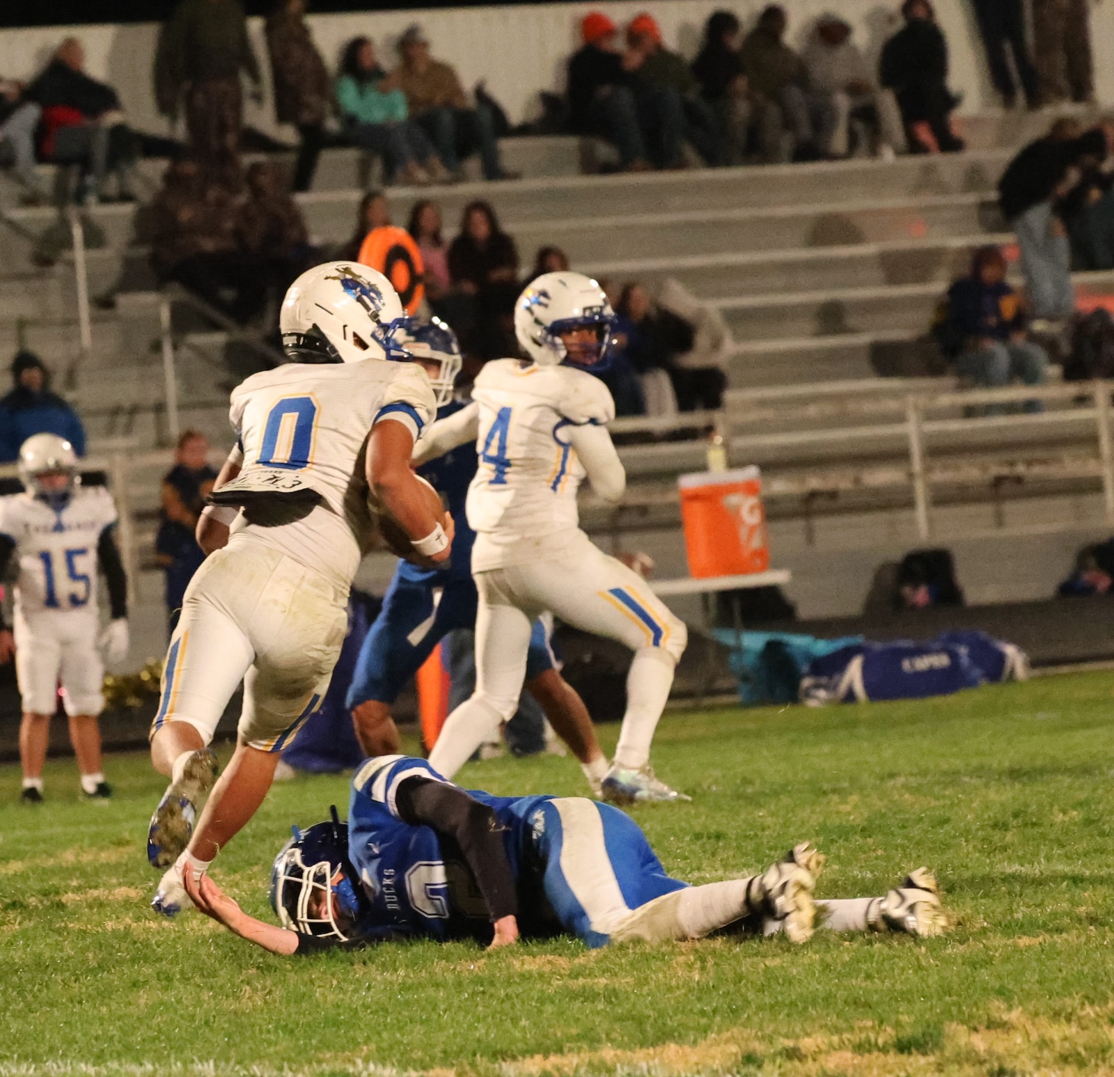 Football players in white uniforms with blue numbers chase a blue-clad opponent; a tackle happens on the grass with bleachers and spectators in the background.