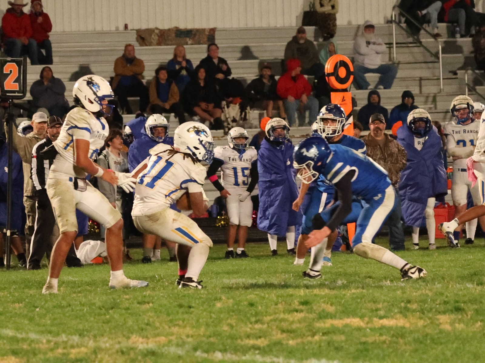 Football players in white and blue uniforms clash on a grassy field as spectators watch from the bleachers in the background.
