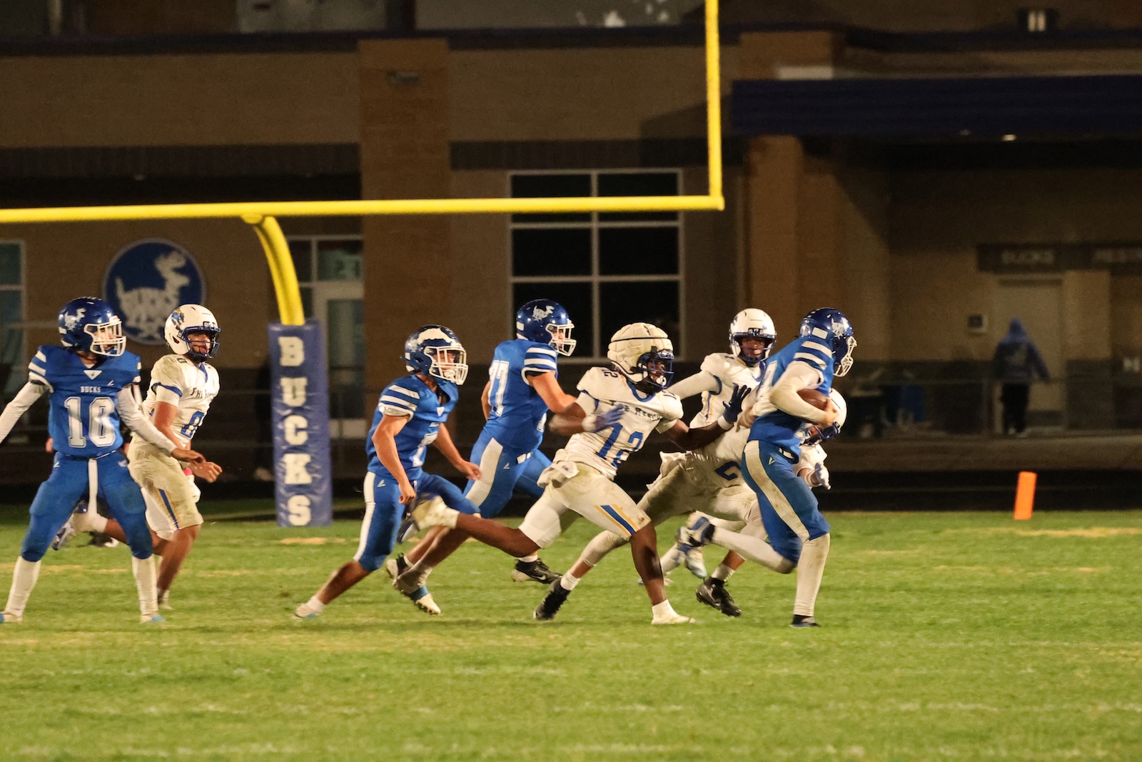 Nighttime high school football game: a blue-uniformed runner carries the ball as white-uniformed players chase him near the end zone.
