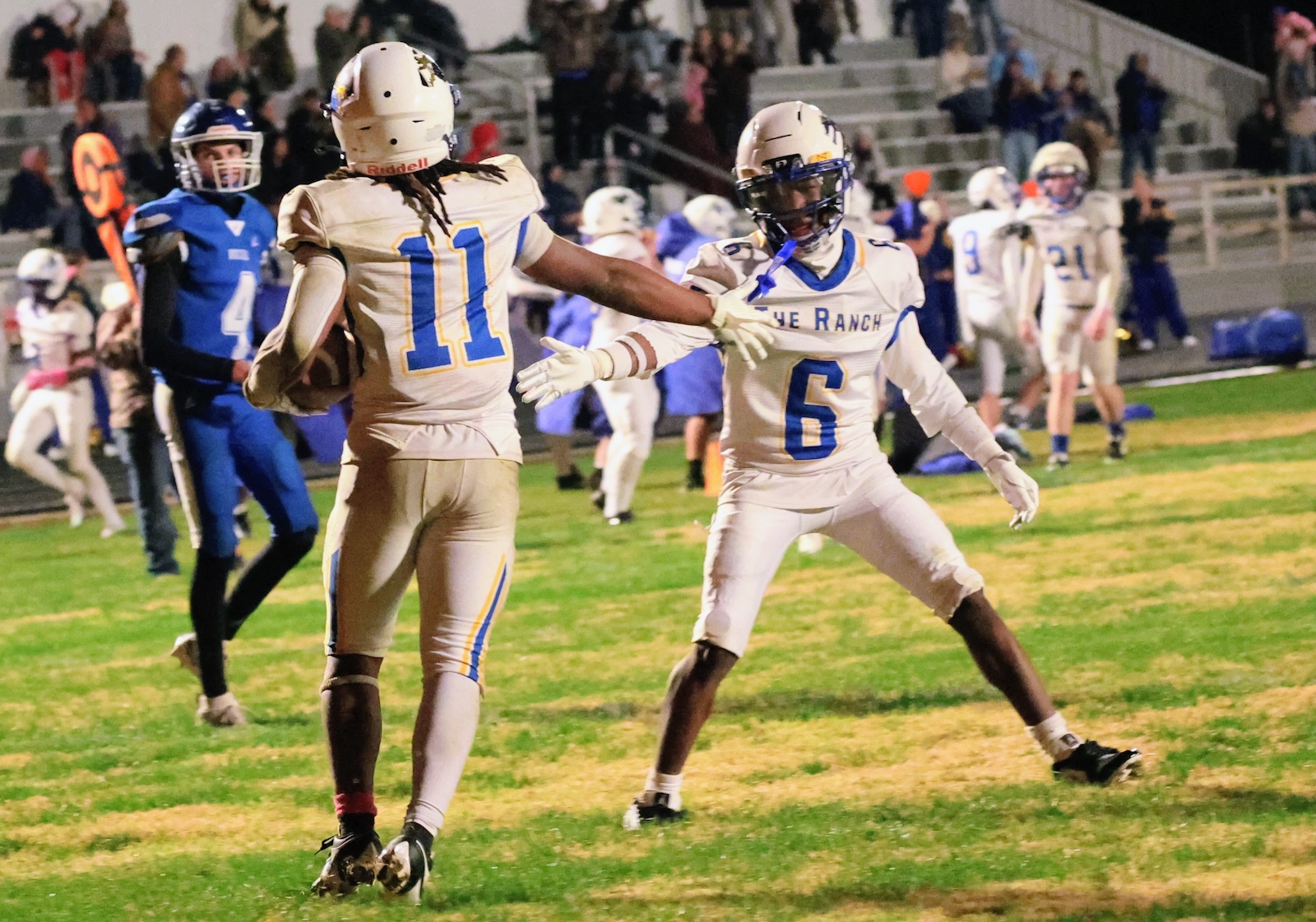 Football players in gold uniforms on a grassy field; #11 carries the ball while teammate #6 extends an arm in support amid a blue-clad defender and onlookers in the stands.