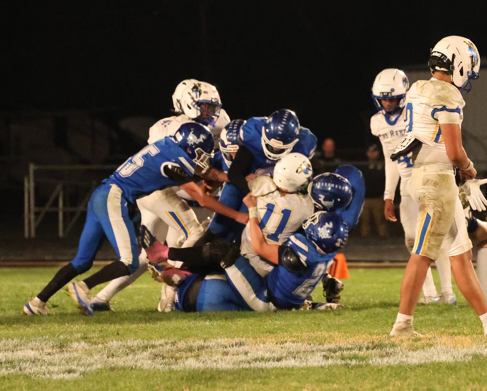 Group of football players in blue and white pile up on the grass during a night game, tackling the ball carrier.
