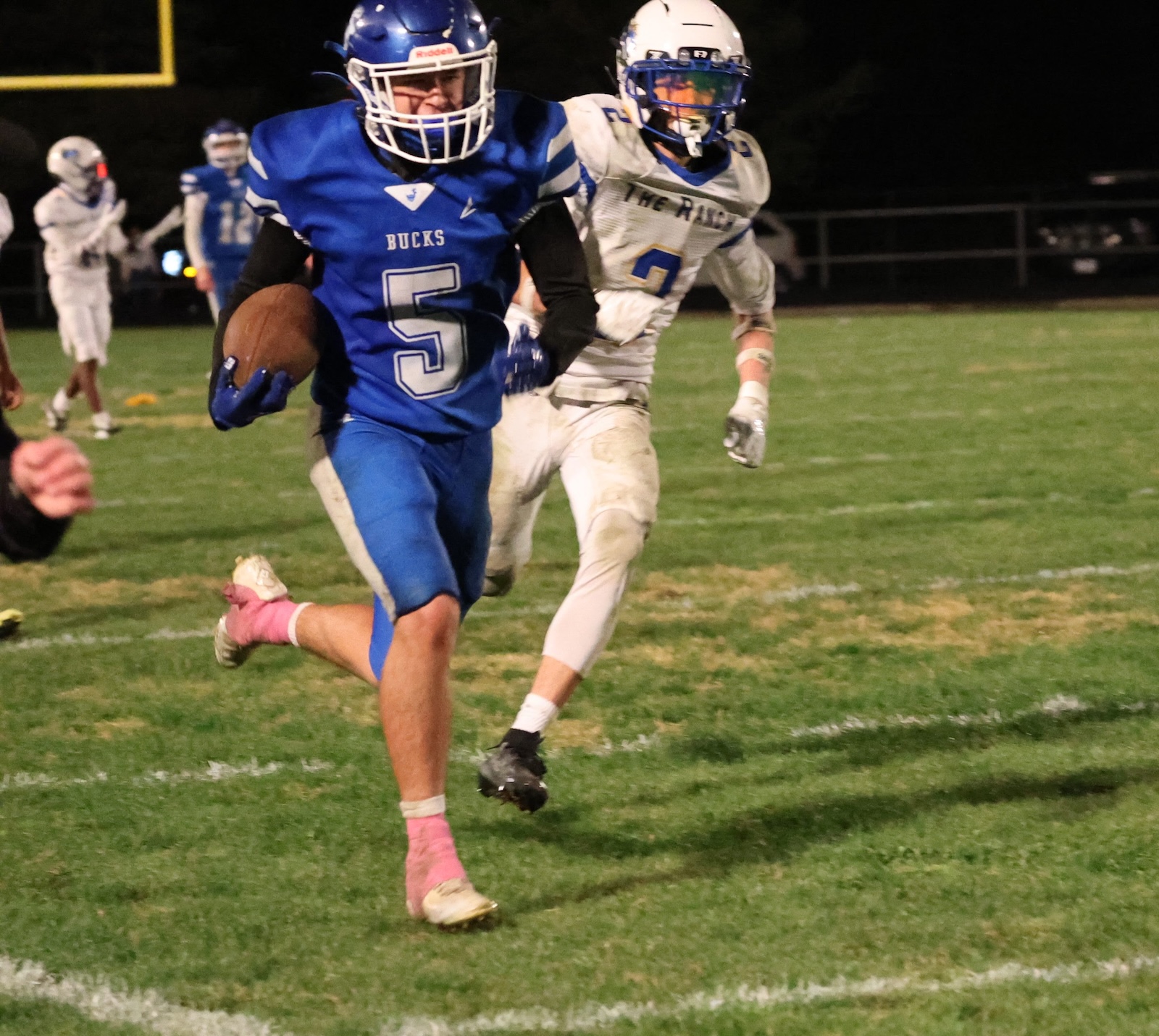 Young football player in blue uniform (No. 5) runs with ball while pursued on a night game field.