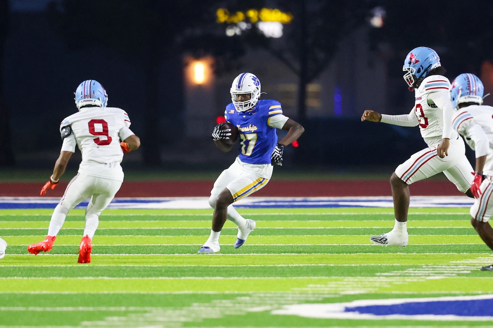 Seabolt Chase-18708 | Press Pass Sports Football player in blue and yellow jersey runs with the ball as white-uniformed defenders close in during a night game on a bright, green field.