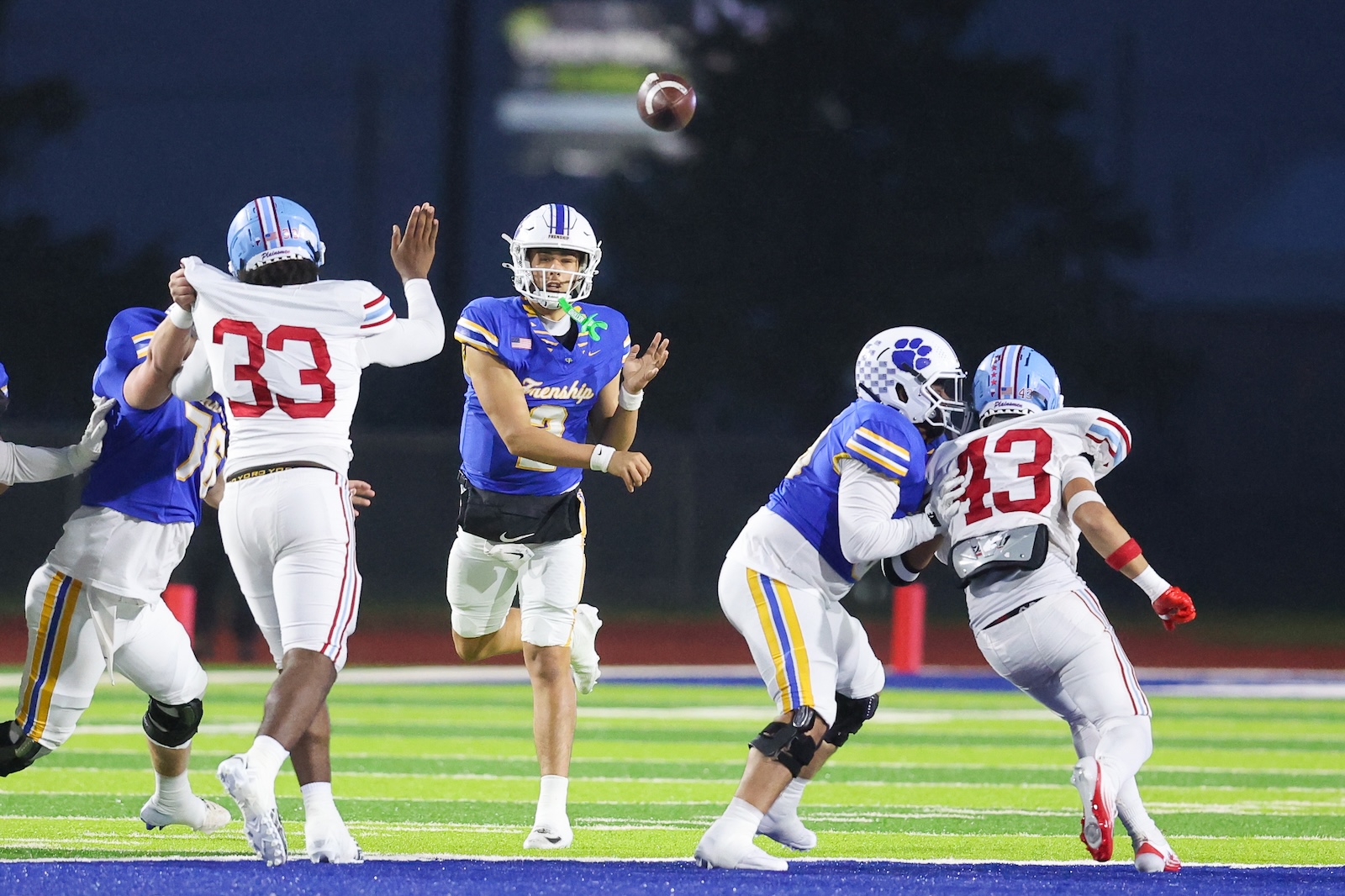 Seabolt Chase-18740 | Press Pass Sports Quarterback in blue throws a pass as teammates and opponents converge on a brightly lit football field at night.
