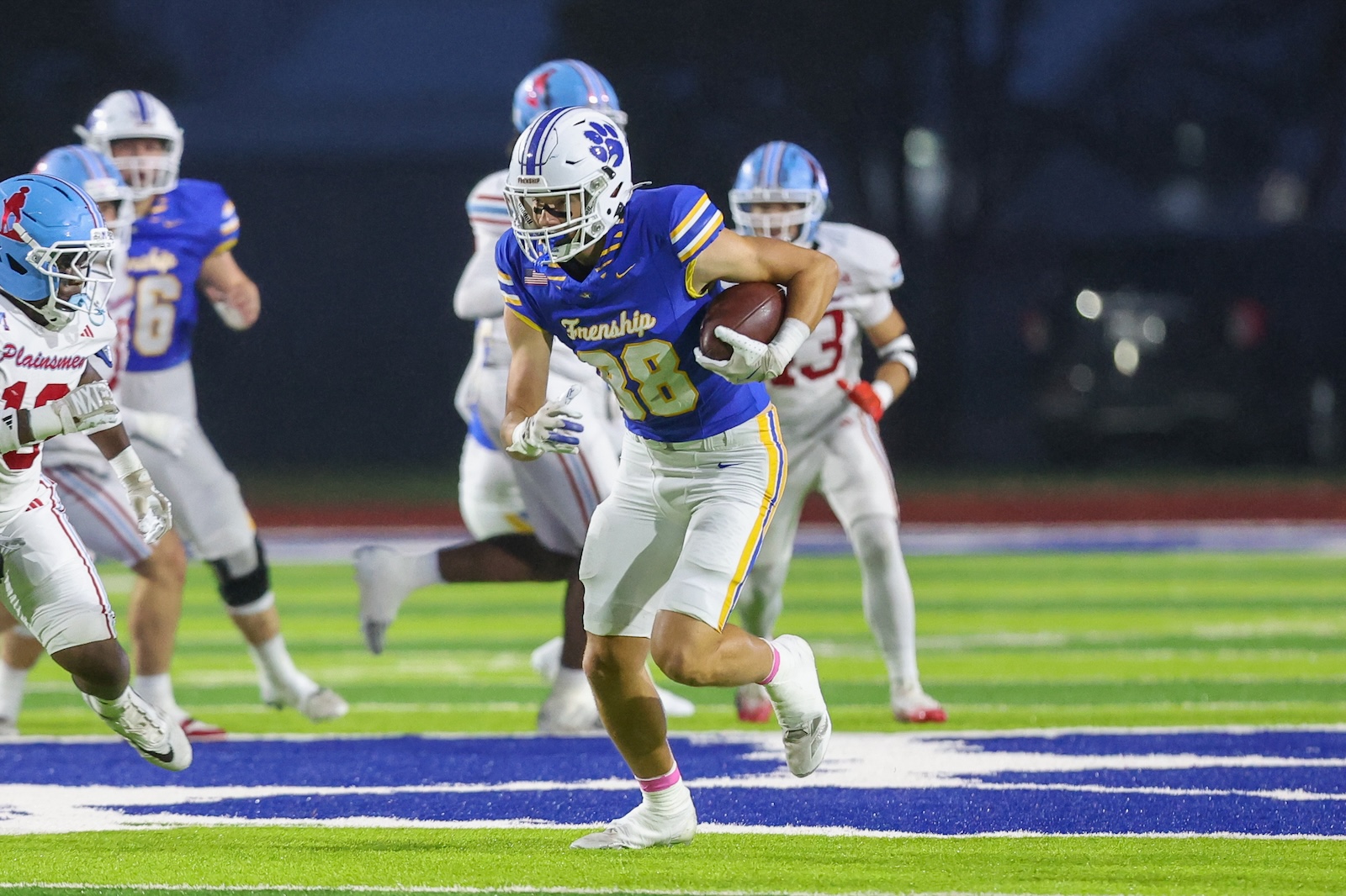 Seabolt Chase-18750 | Press Pass Sports Running back in a blue and yellow football uniform grips the ball and sprints down the field under stadium lights, pursued by opponents.