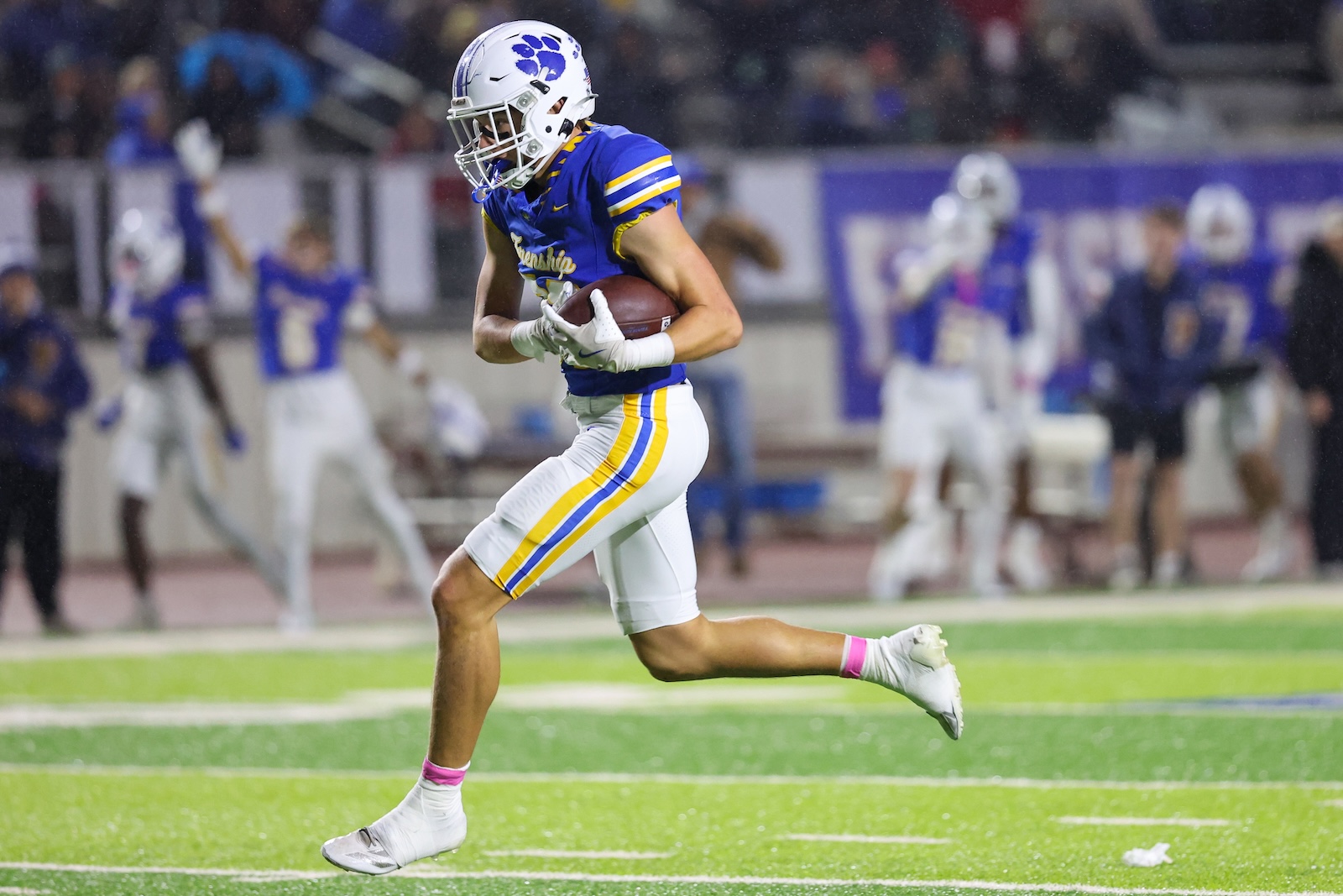Seabolt Chase-18810 | Press Pass Sports Football player in blue and gold uniform running with the ball on a bright green field, stadium and teammates in the background.