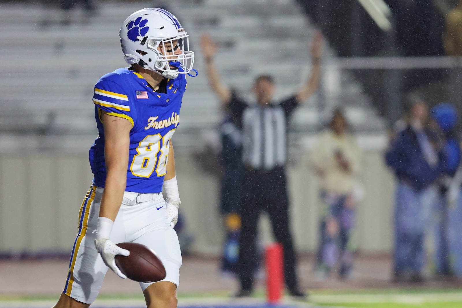 Seabolt Chase-18847 | Press Pass Sports Football player in blue and yellow jersey (#84) holds the ball and runs, with a referee and blurred crowd in the background in the rain
