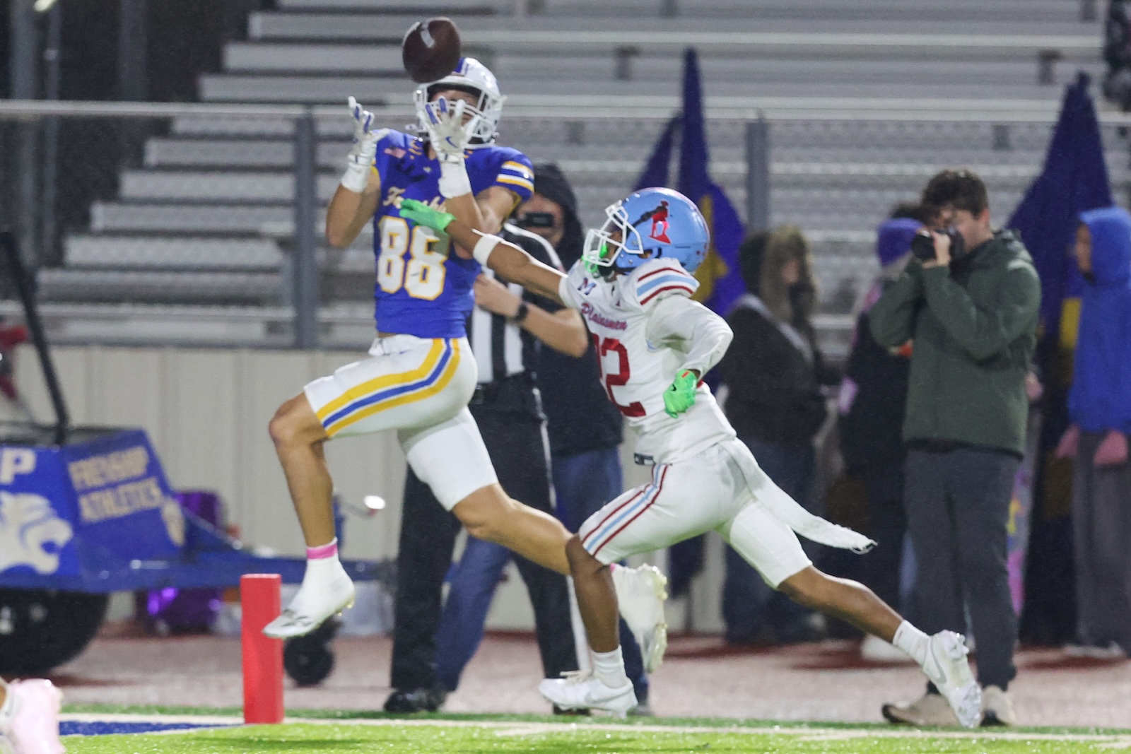 Seabolt Chase-19261 | Press Pass Sports Two football players jump for the ball near the sideline as a pass is contested, blue and gold team vs white uniforms with red numbers, crowd in background.