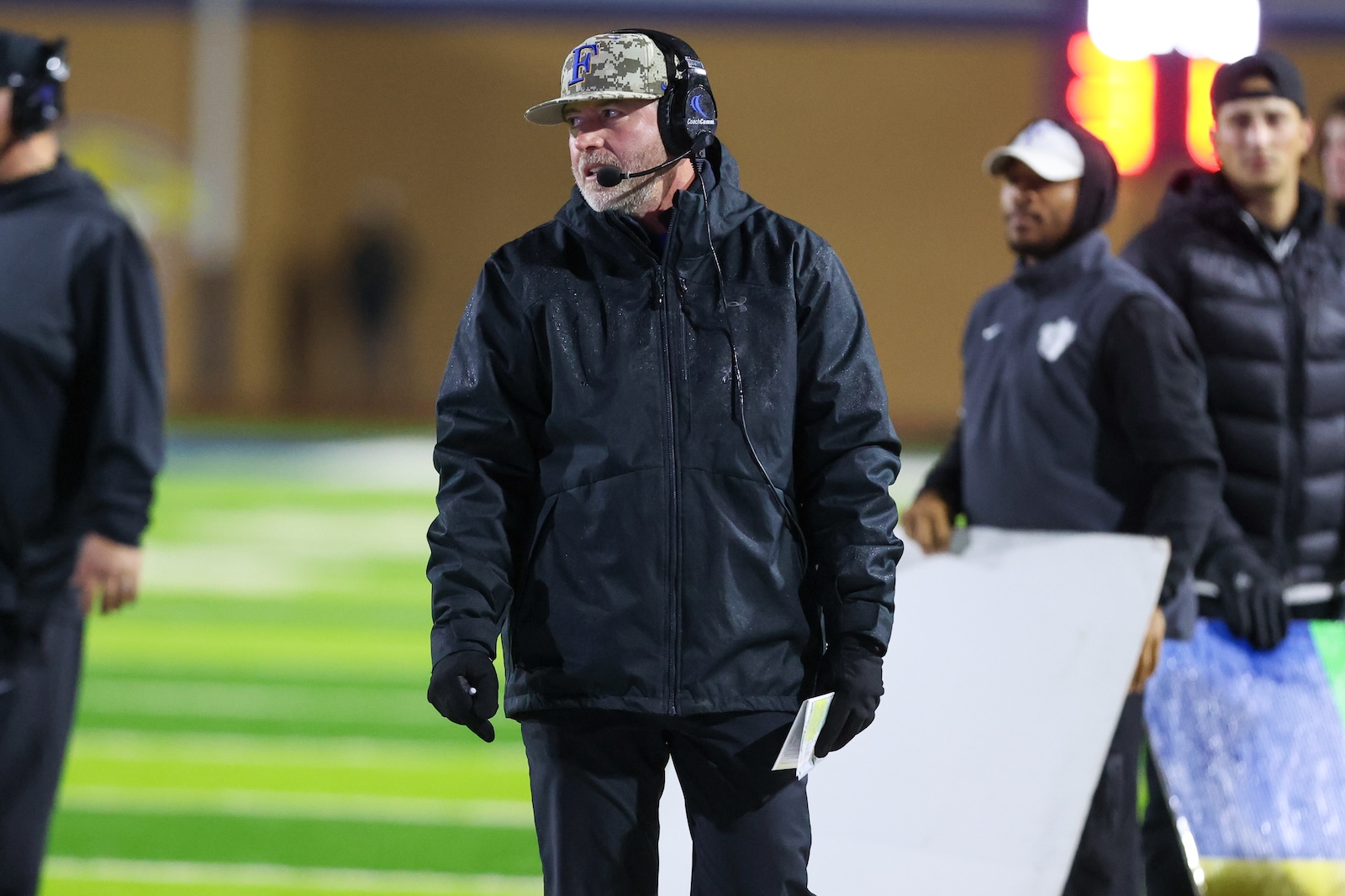 Seabolt Chase-19467 | Press Pass Sports Coach wearing a camo cap and headset on the sideline of a football field, holding a clipboard in rainy weather