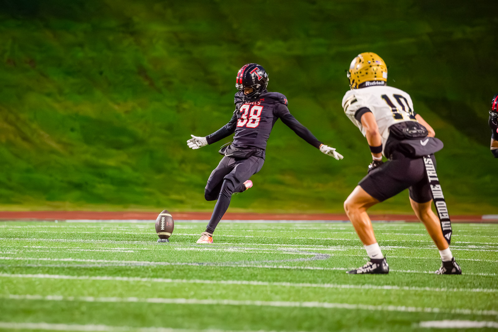Football game at night: kicker in black #38 about to kick the ball as an opponent in white #10 closes in on the field.