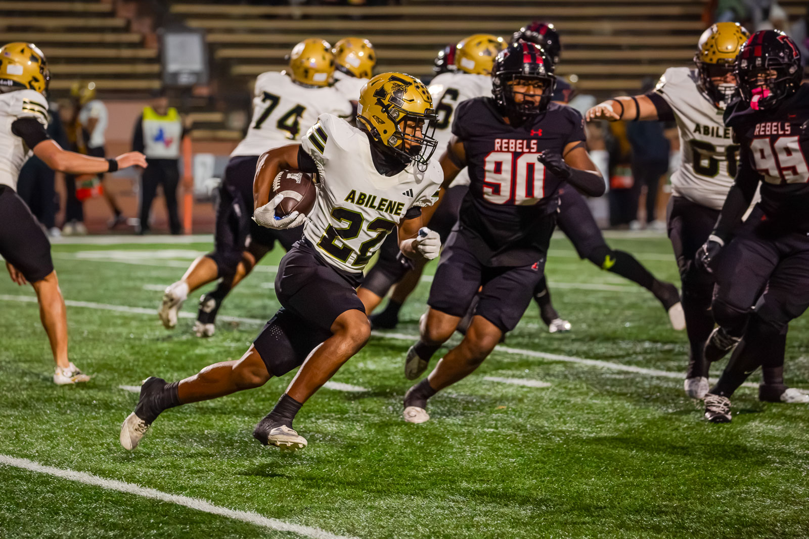Abilene football player #22 in white jersey runs with the ball during a night game, pursued by opponents on a green turf field.
