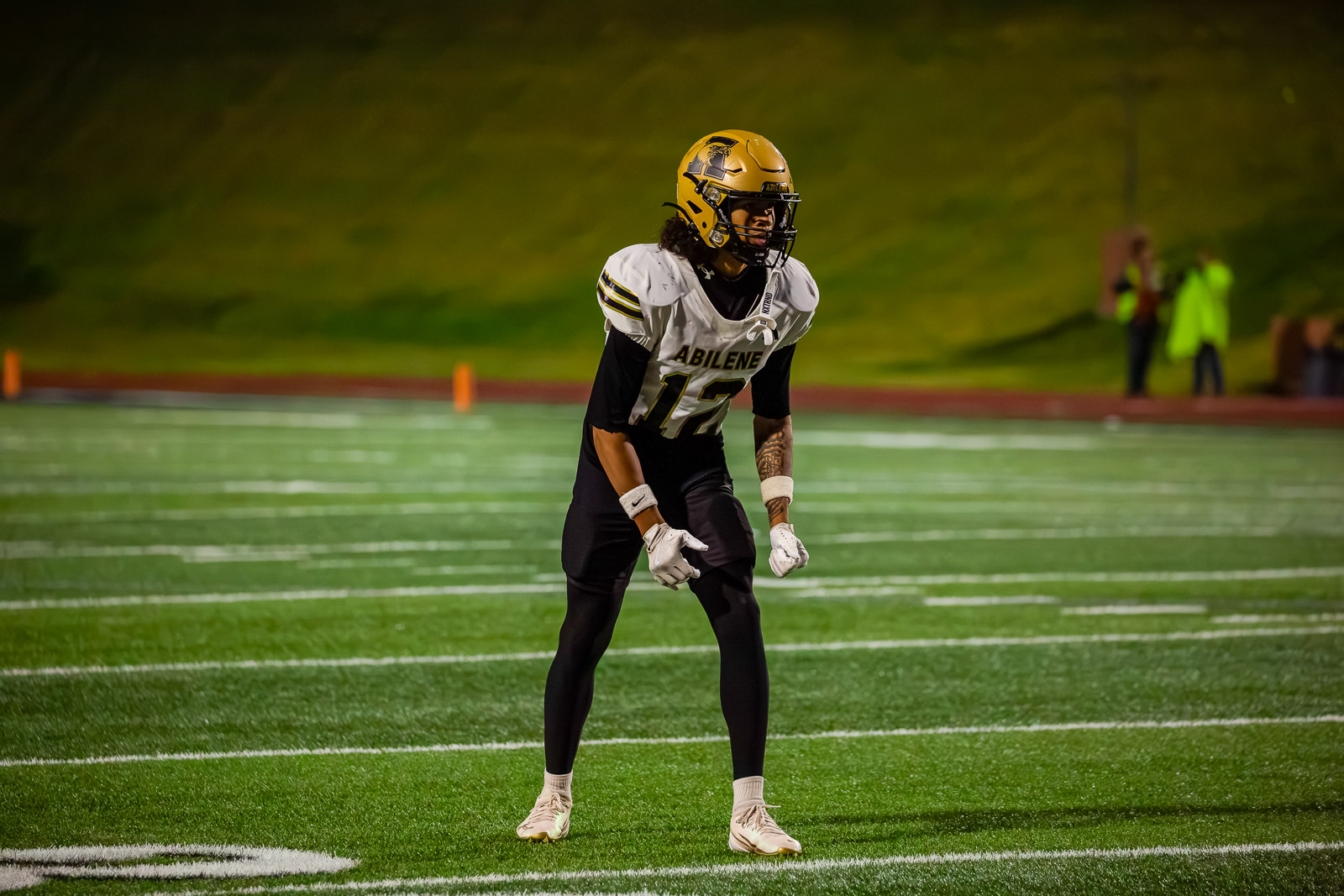 Football player in gold helmet and white Abilene jersey number 12, crouched ready on a green turf field at night.