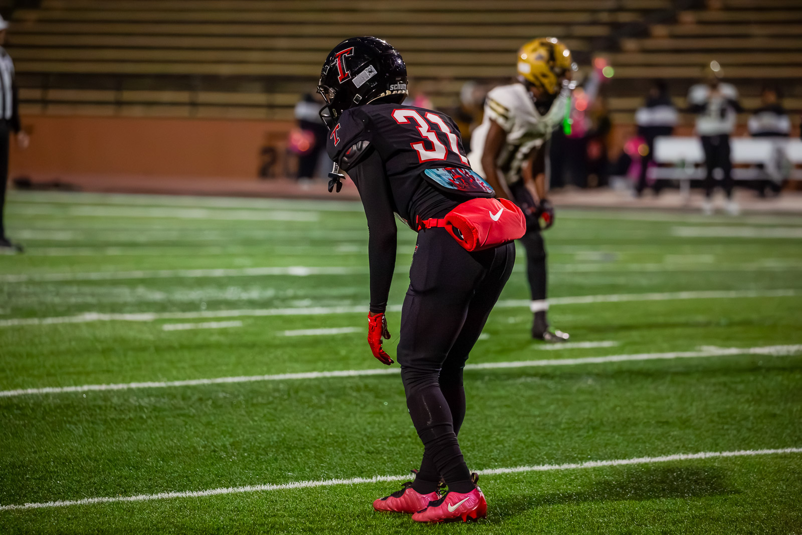 Football player in black uniform with number 31, crouched in stance on a green turf field, red gloves and waist pack visible