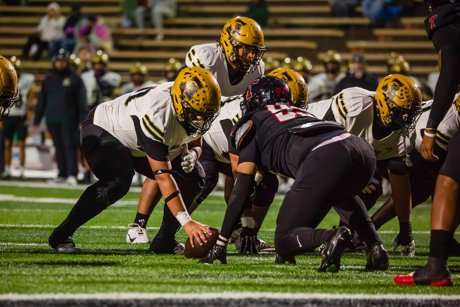 Offensive linemen in white jerseys and gold helmets crouch at the line of scrimmage as the ball is snapped.