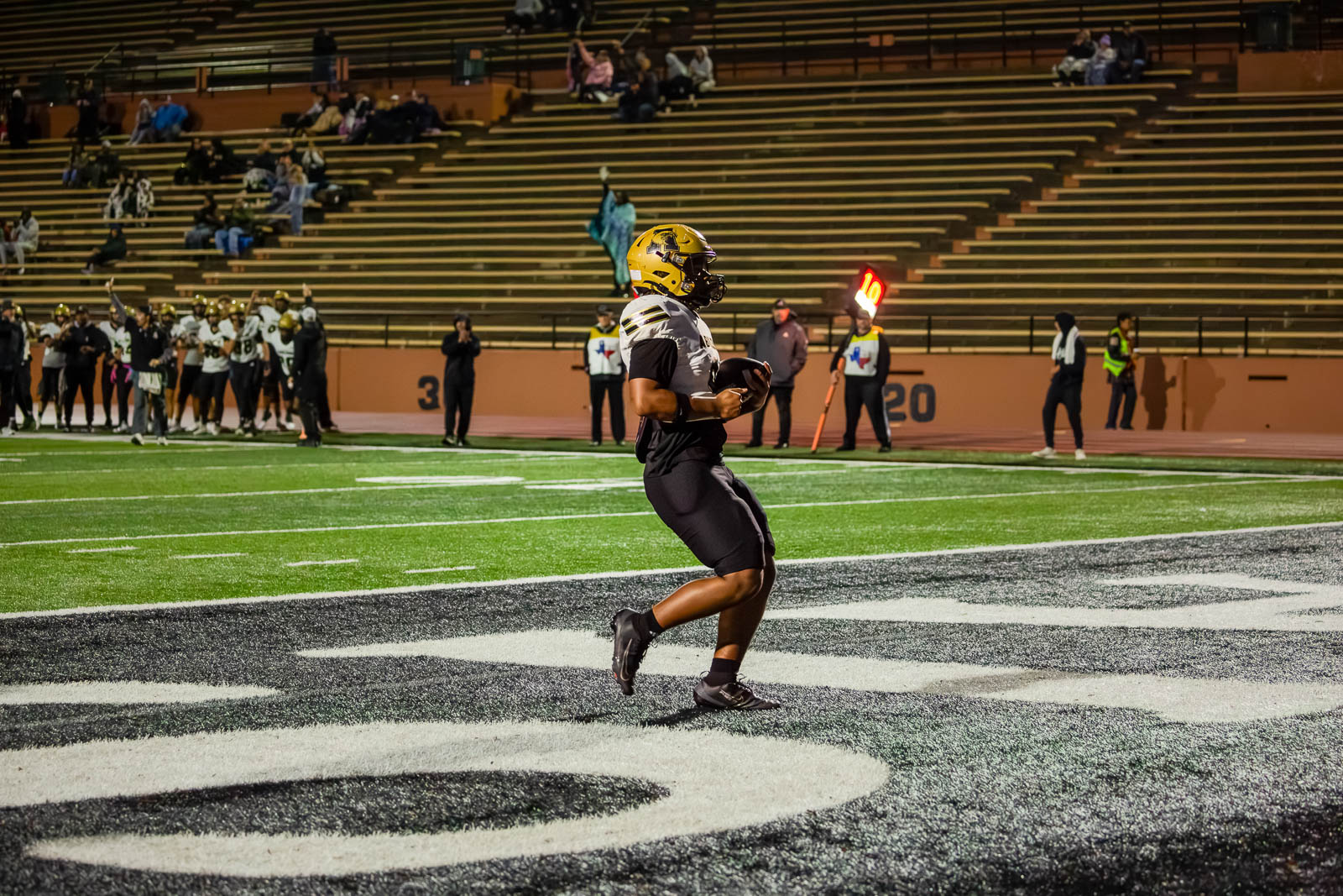 Football player in a black and gold uniform runs along the sideline with his helmet on, holding the ball.