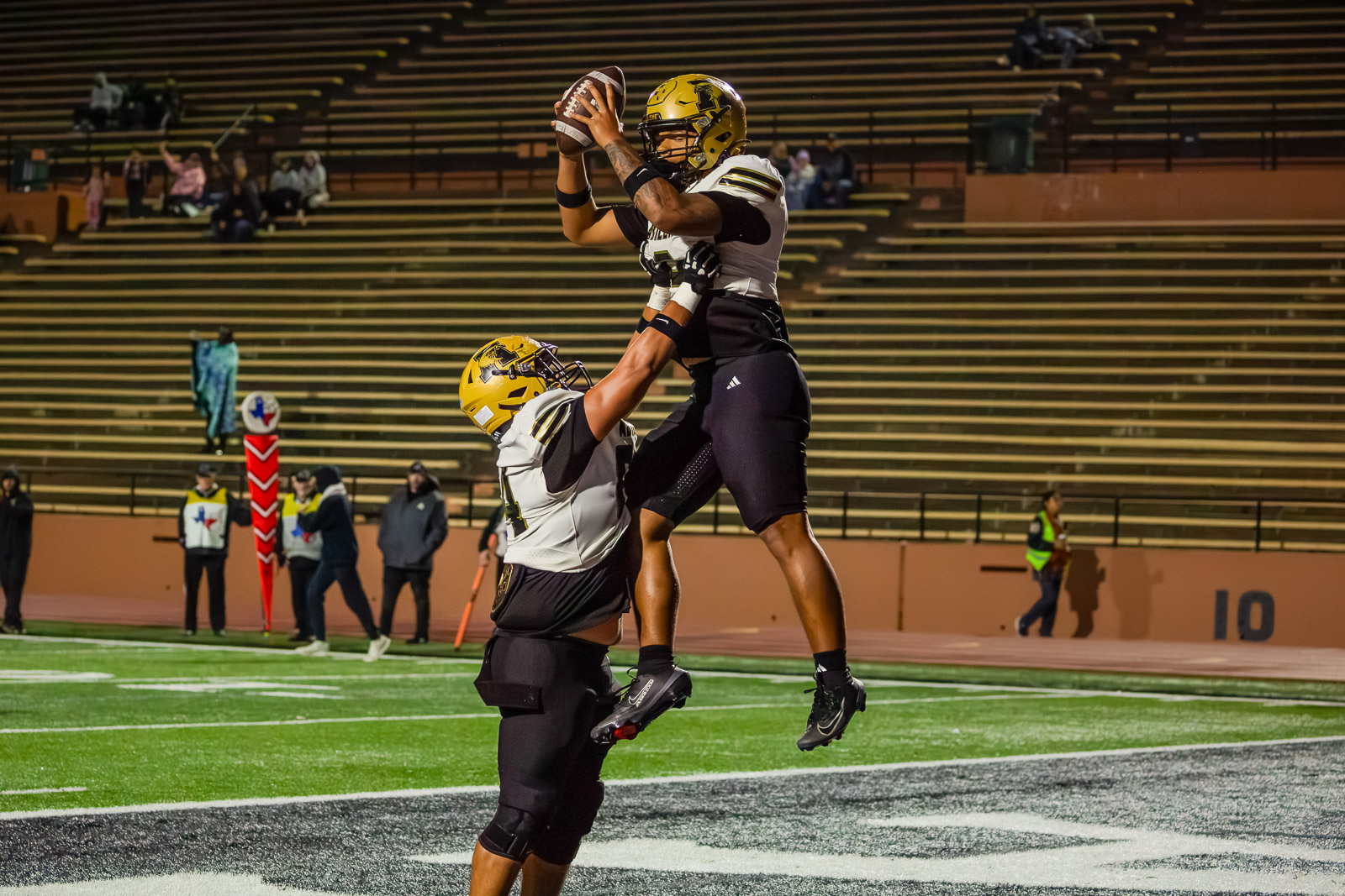 Football players in yellow helmets celebrate a leaping catch near the end zone as a teammate lifts him up under stadium lights