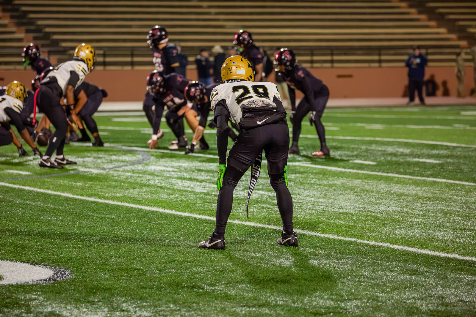 Football player wearing a gold helmet and white jersey with #29 stands ready at the line of scrimmage on a green turf field at night, gloves glowing green.