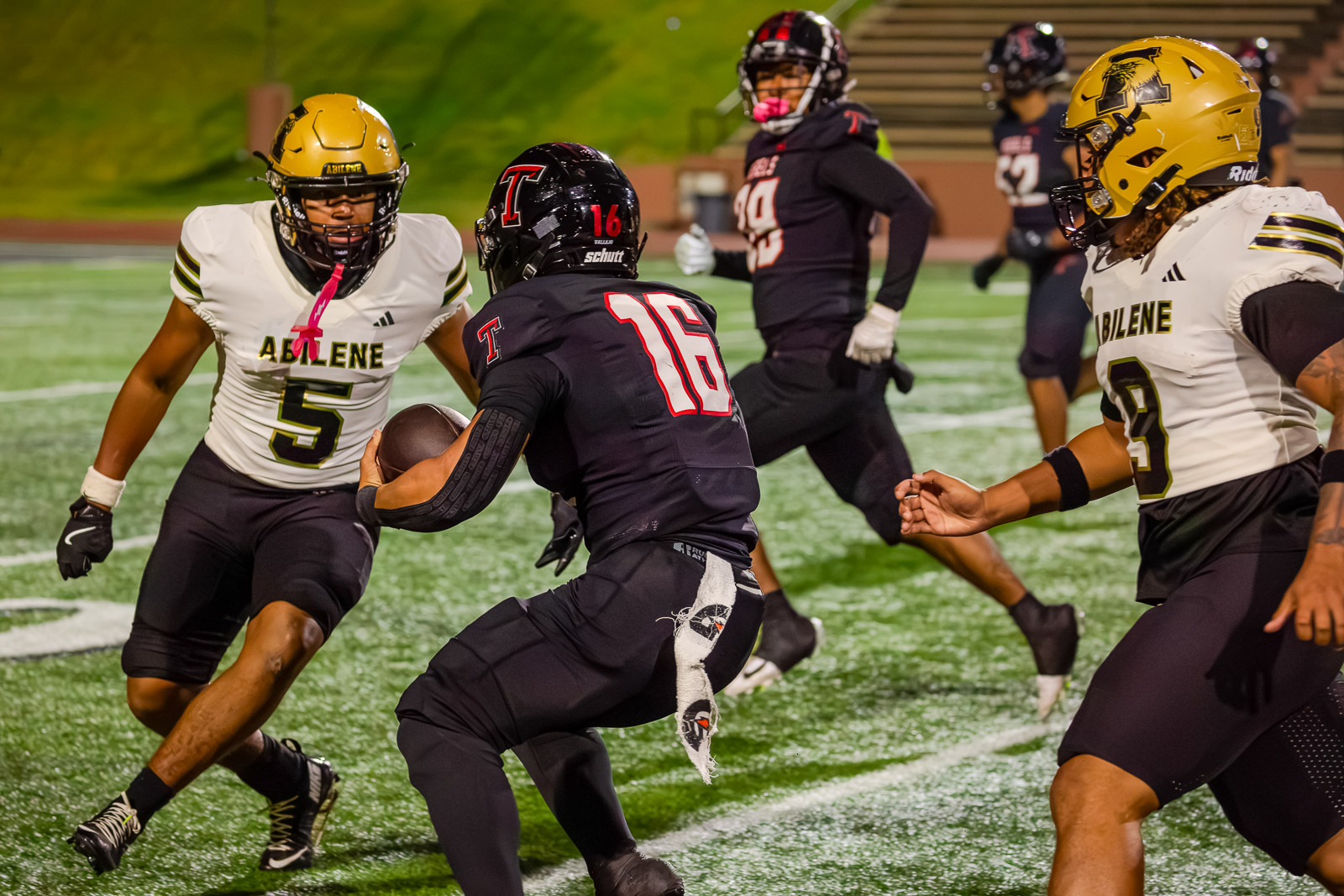 Football action: a ball carrier in black #16 cuts through two Abilene defenders in white and gold uniforms on a green turf field at night.