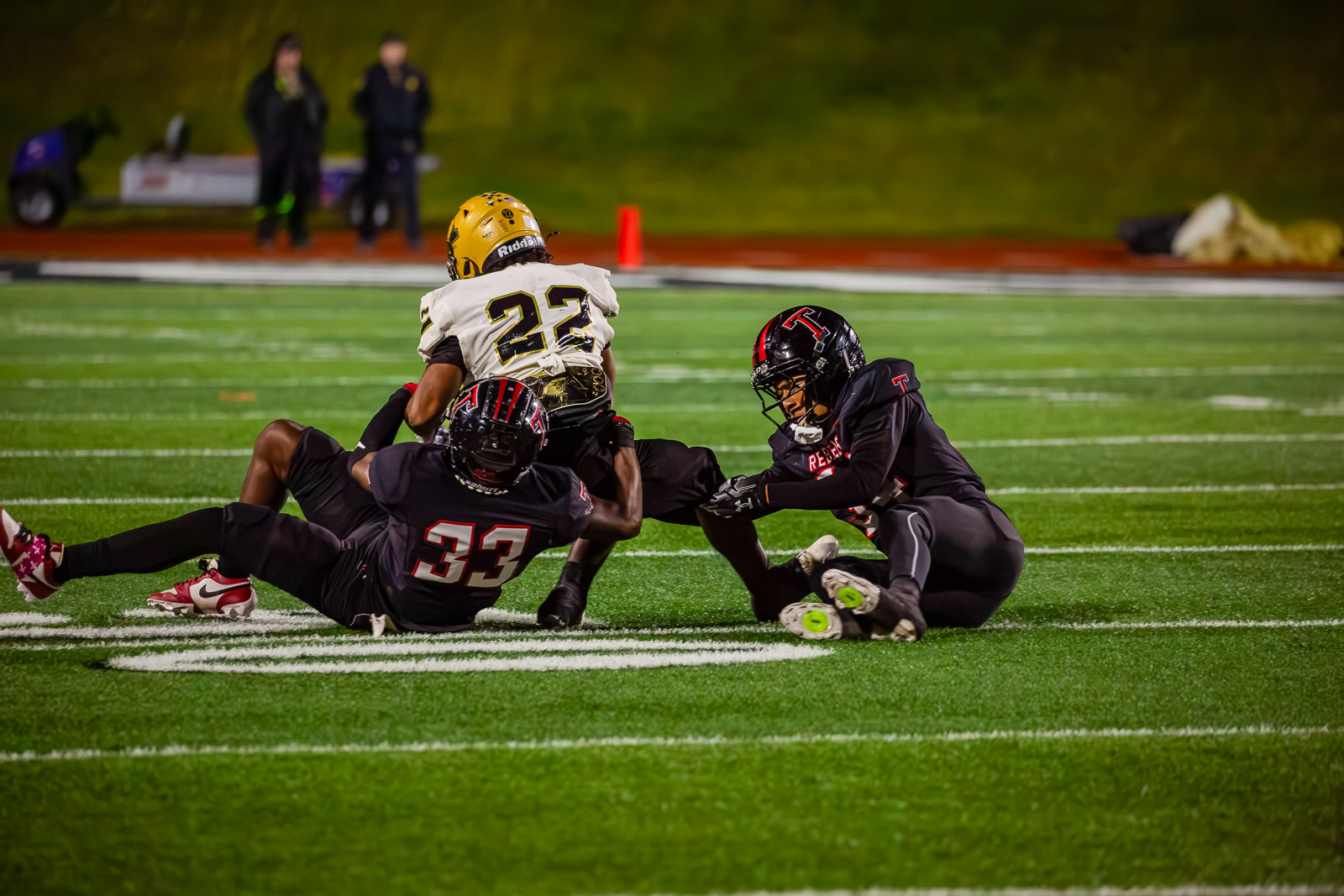 Football players tackle during a nighttime game; a white-jersey runner (#23) is grabbed by two black-jersey defenders on a green turf field.