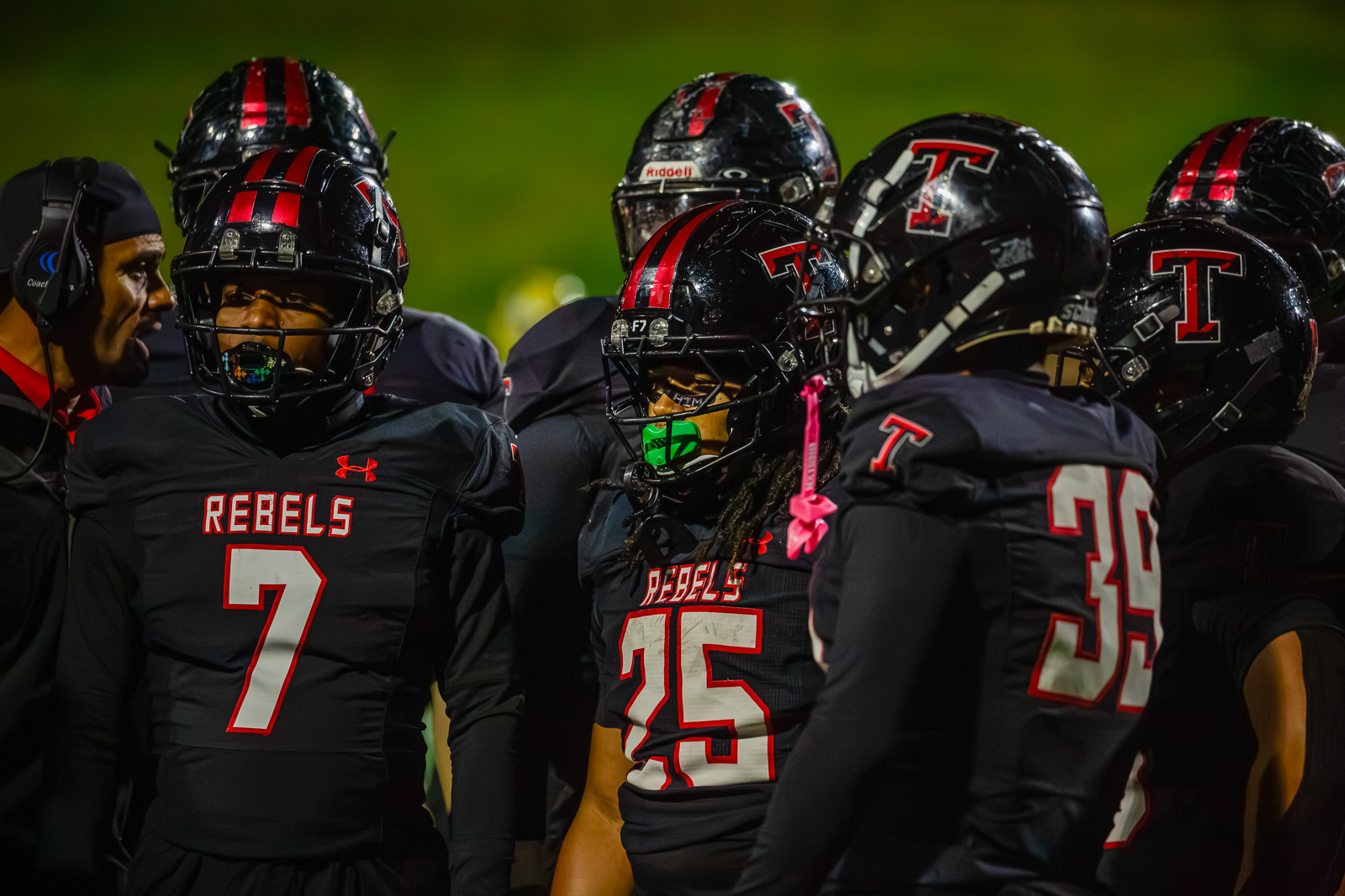 Group of football players in black jerseys with red numbers and the word 'REBELS' gathered on a darkened field at night.