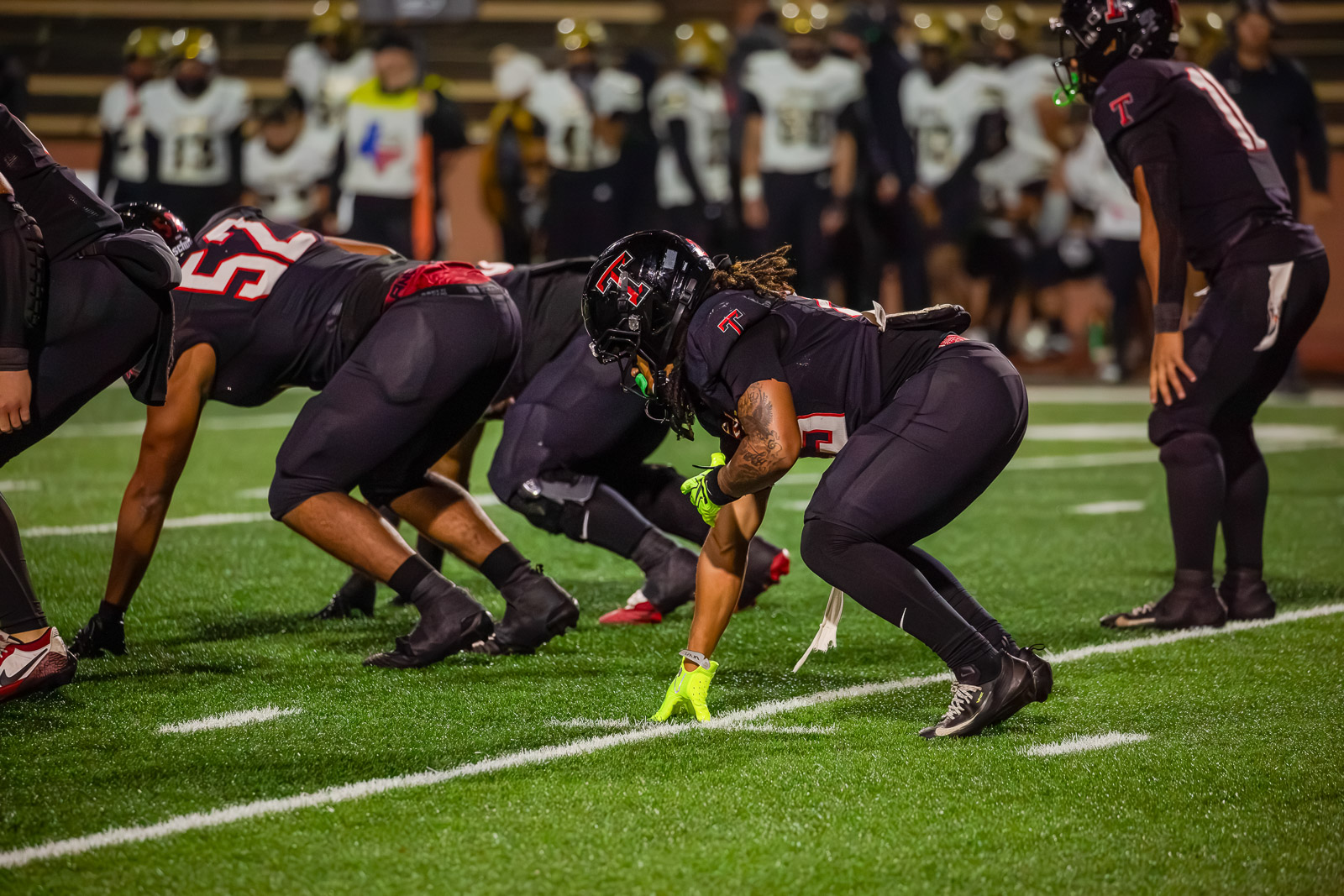 American football players line up at the line of scrimmage on a green turf field at night, wearing black uniforms and neon gloves.