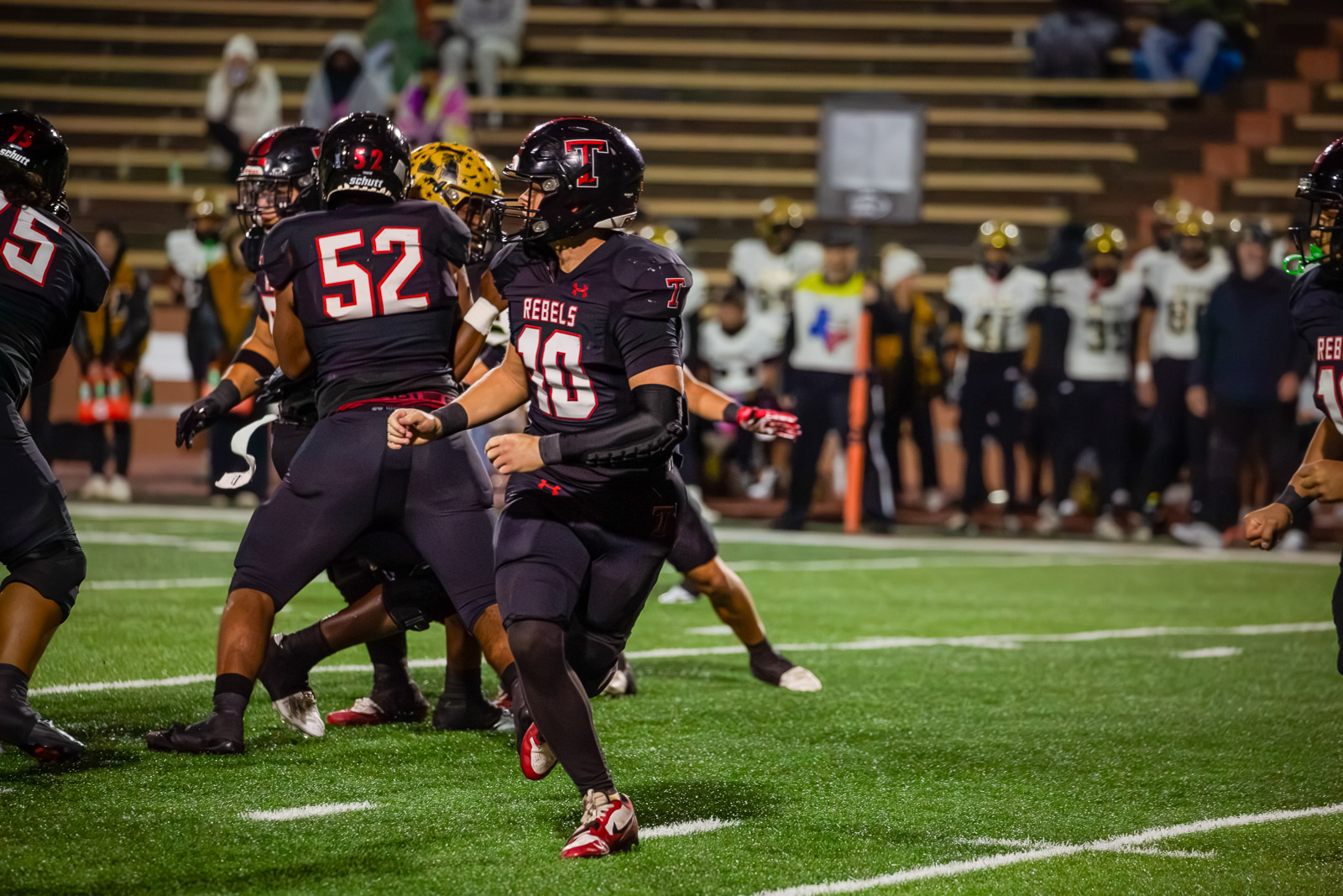 Football players in black Rebels jerseys clash at the line of scrimmage on a bright turf field during a night game. (number 40 visible)