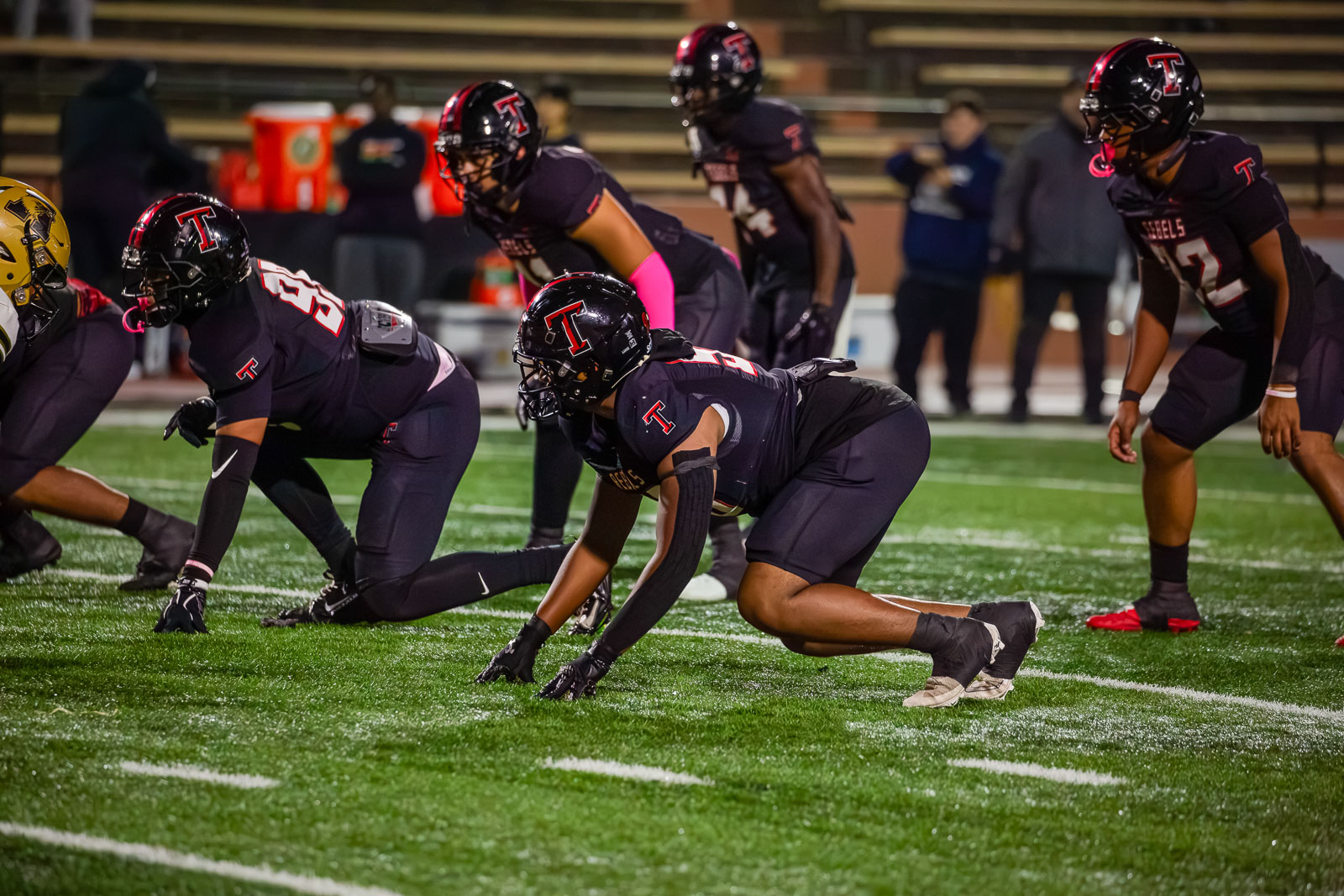 Football players in dark uniforms crouch at the line of scrimmage on a turf field during a nighttime game.