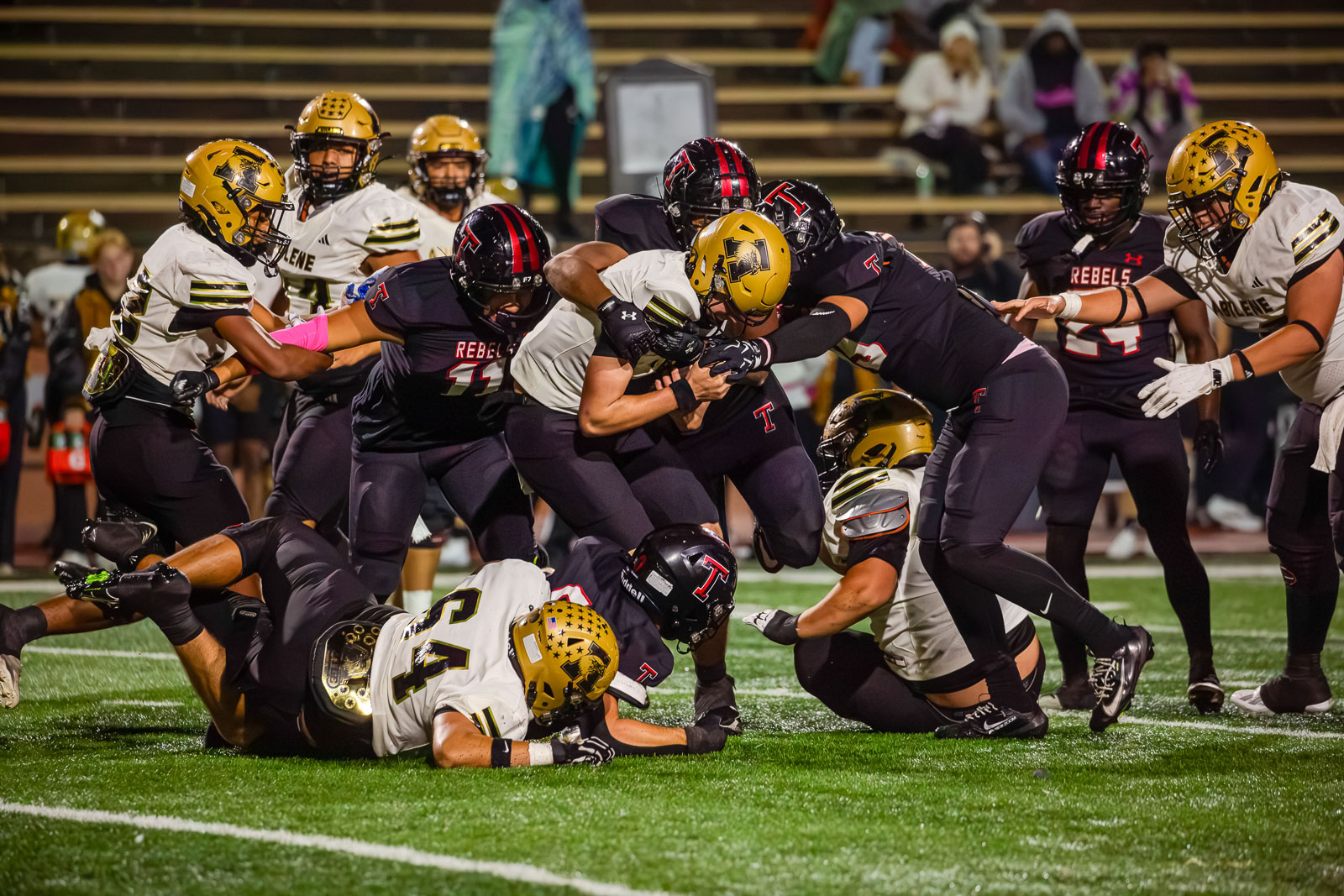 Football players in black and gold jerseys collide in a crowded tackle on the field during a night game at the stadium.
