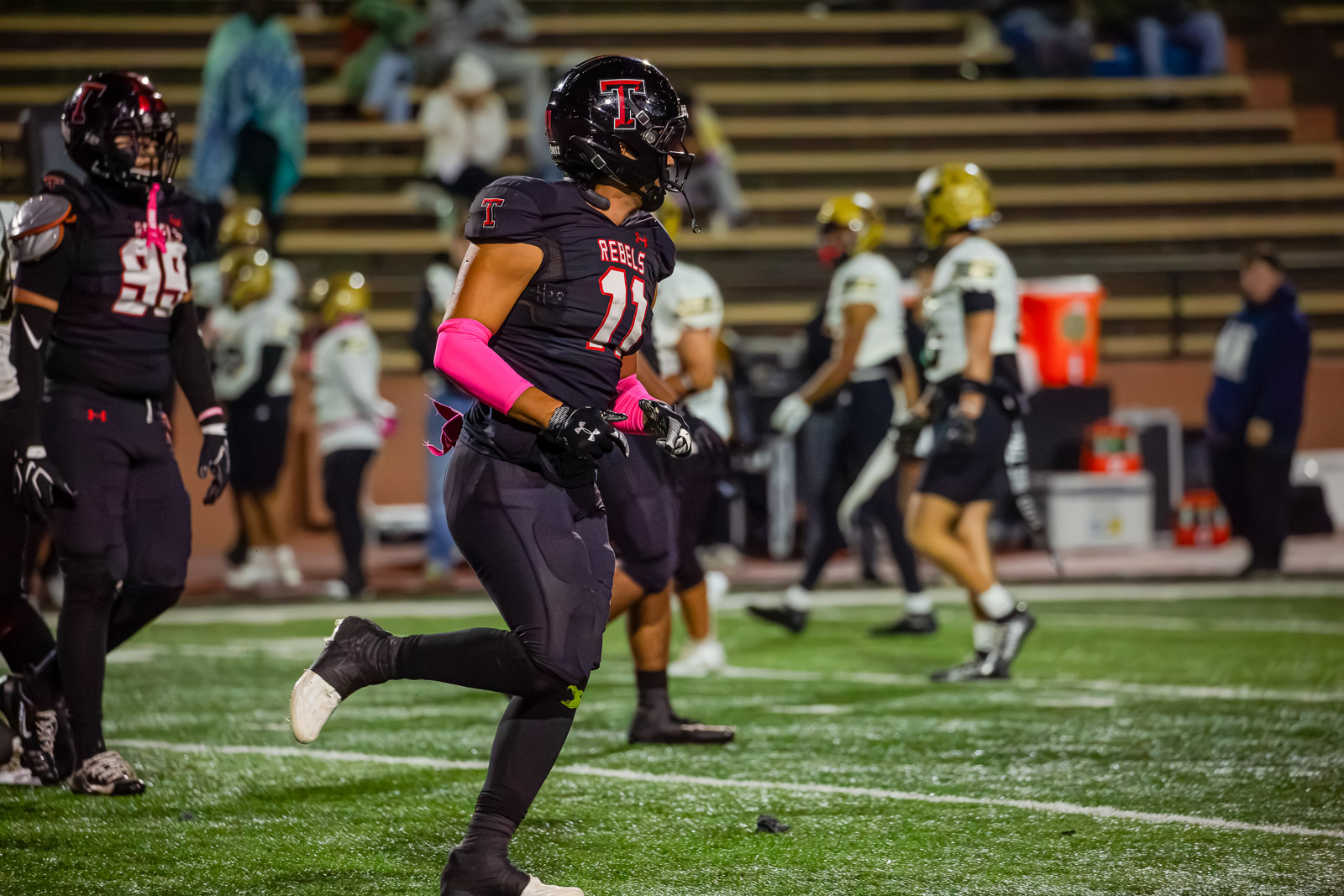 Football player in a black Rebels uniform (number 11) runs on the turf field with pink arm sleeves during a game, opponents in white/gold uniforms in the background.