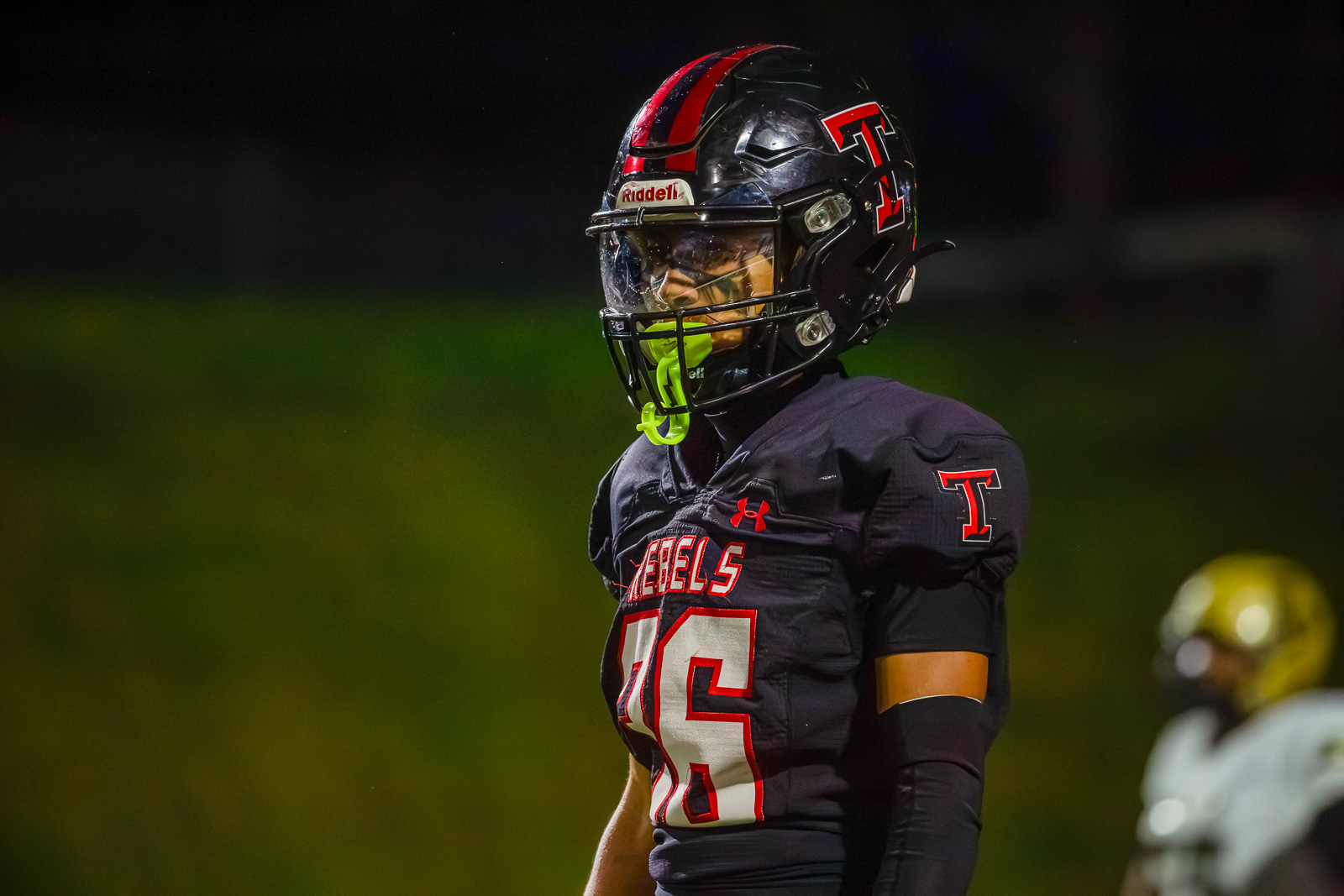 Football player in a black jersey with red trim and number 25, wearing a helmet, standing on a dimly lit field at night.