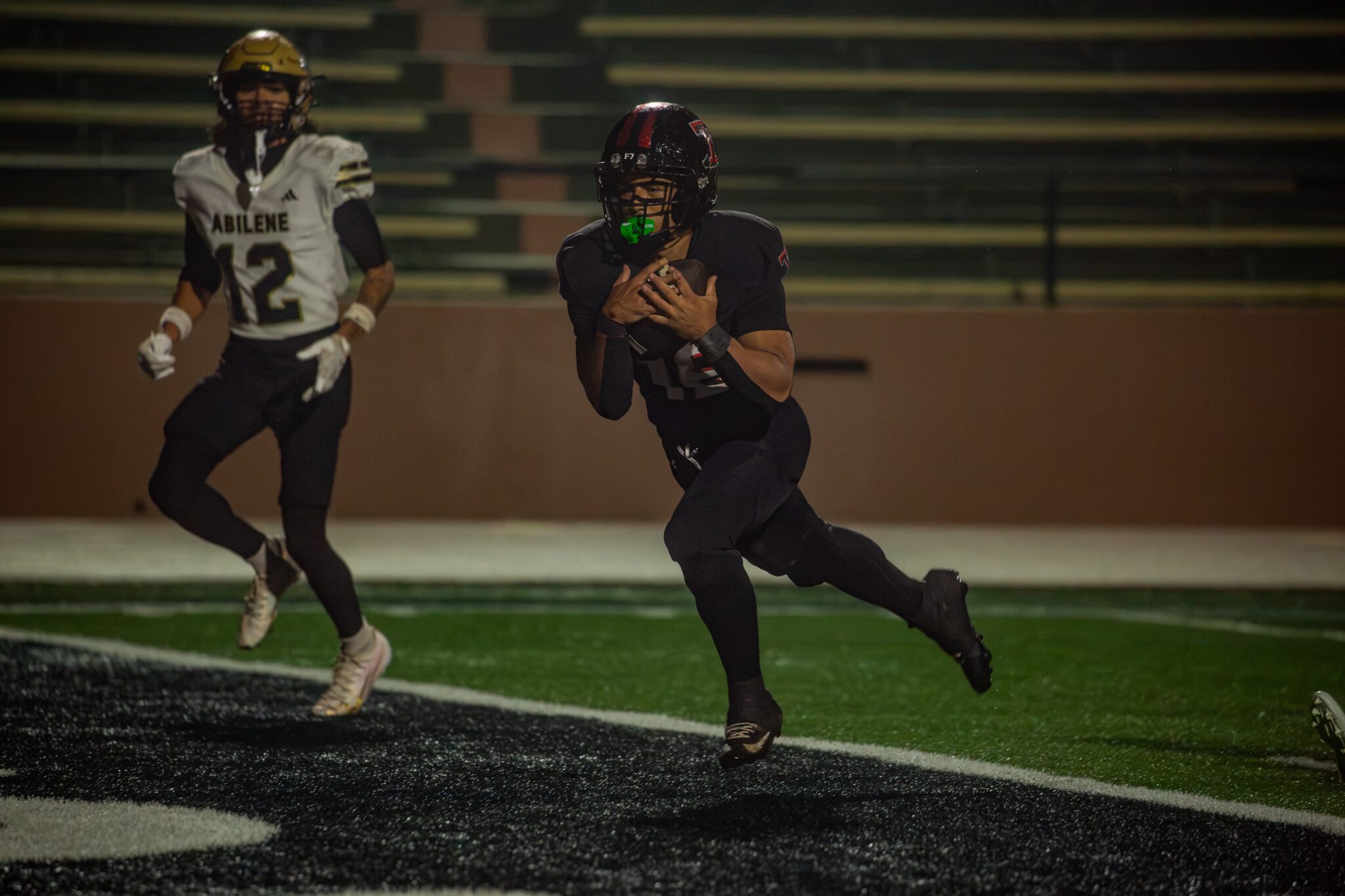 Football player in a black jersey runs with the ball during a night game, blurred stadium bleachers in the background, players in the foreground.