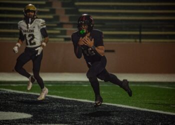 IMG_4354 | Press Pass Sports Football player in a black jersey runs with the ball during a night game, blurred stadium bleachers in the background, players in the foreground.