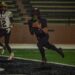 IMG_4354 | Press Pass Sports Football player in a black jersey runs with the ball during a night game, blurred stadium bleachers in the background, players in the foreground.